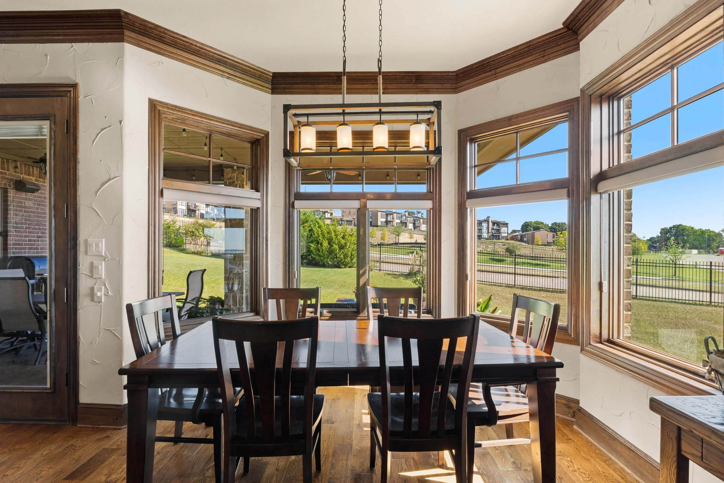 Dining room with large windows, wooden trim, and a wooden table with six chairs. A chandelier hangs above the table, and the outside view includes grass, trees, and modern buildings.