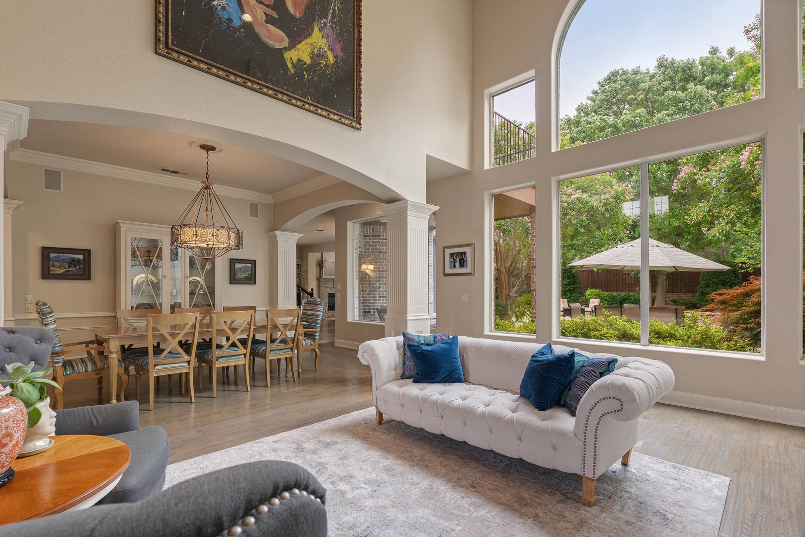 Living room with high vaulted ceiling, large windows showing a backyard with trees and patio, white tufted sofa with blue pillows, dining area with wooden table and striped chairs, and artwork on the walls.