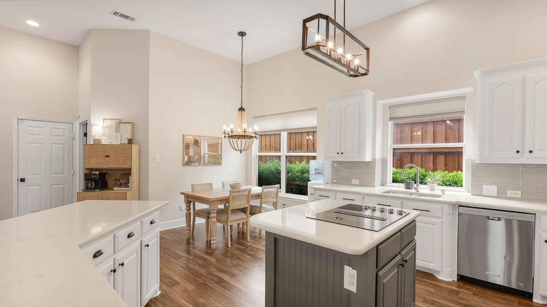 Bright kitchen with white cabinets, a central island, and a dining area with a rustic table and four chairs. Large windows with views of greenery and a wooden fence. Modern lighting fixtures, hardwood flooring, a water dispenser, and a dishwasher.
