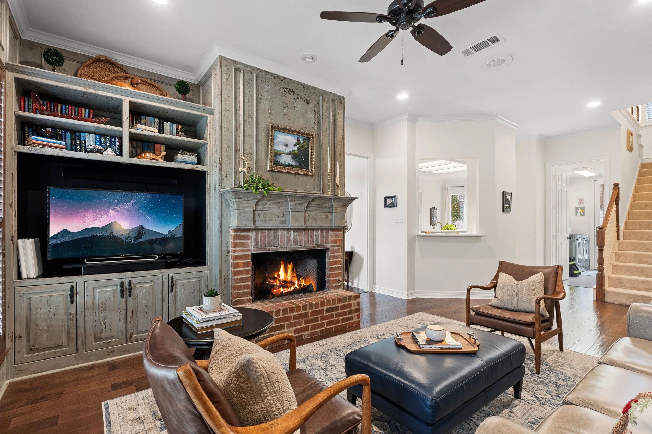Living room with a brick fireplace, a mounted TV above cabinetry, and a ceiling fan. Furniture includes a leather ottoman, a beige sofa, and two chairs with pillows. Decor includes books, plants, and framed artwork.