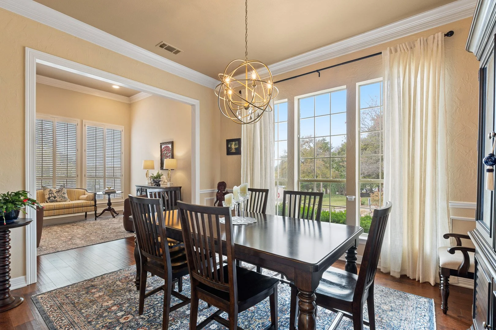 Dining room with a dark wood table and chairs, large windows with curtains, and a chandelier hanging from the ceiling.