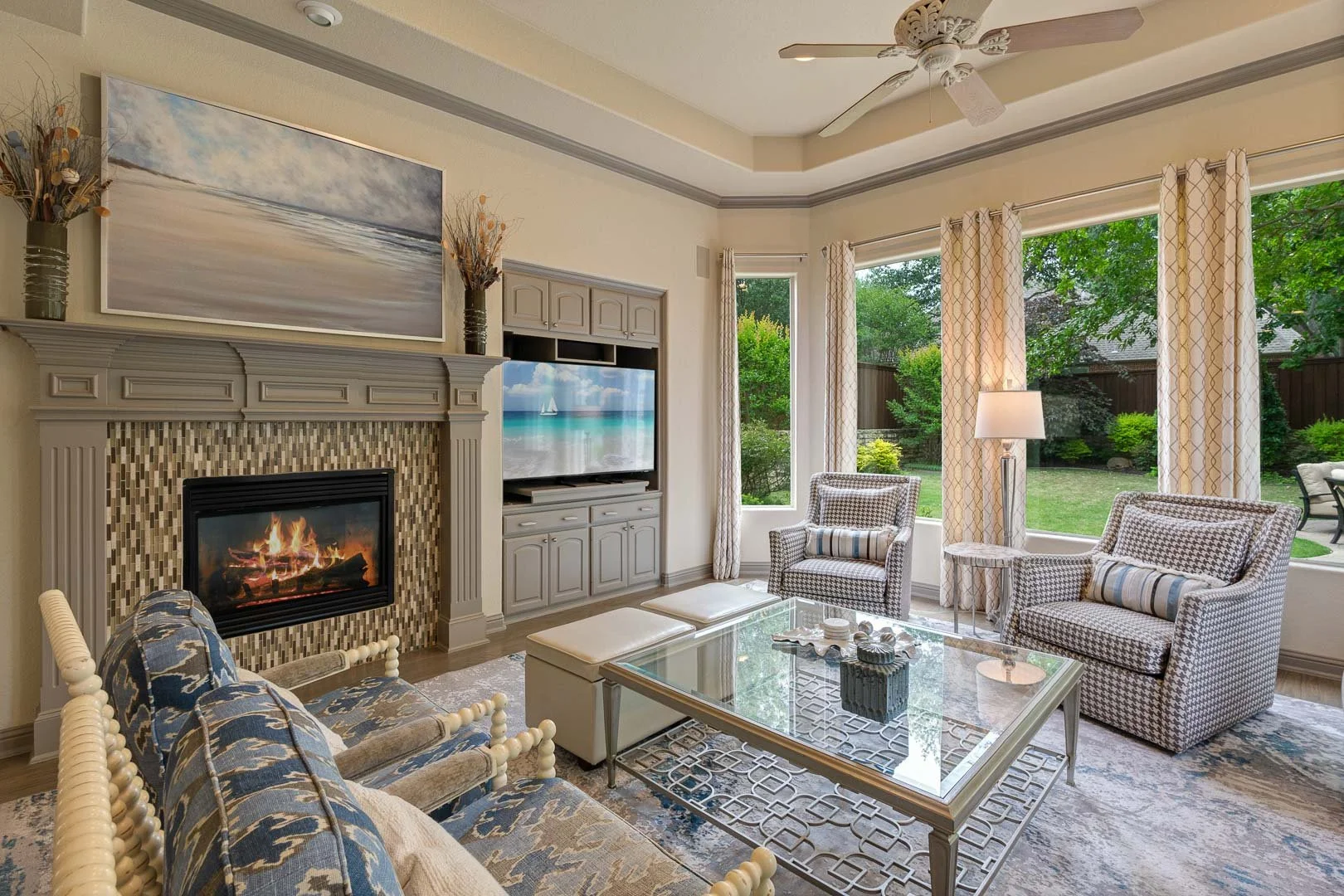 Living room with a fireplace, two armchairs with houndstooth pattern, a glass coffee table, a TV, large windows with curtains, and a ceiling fan.