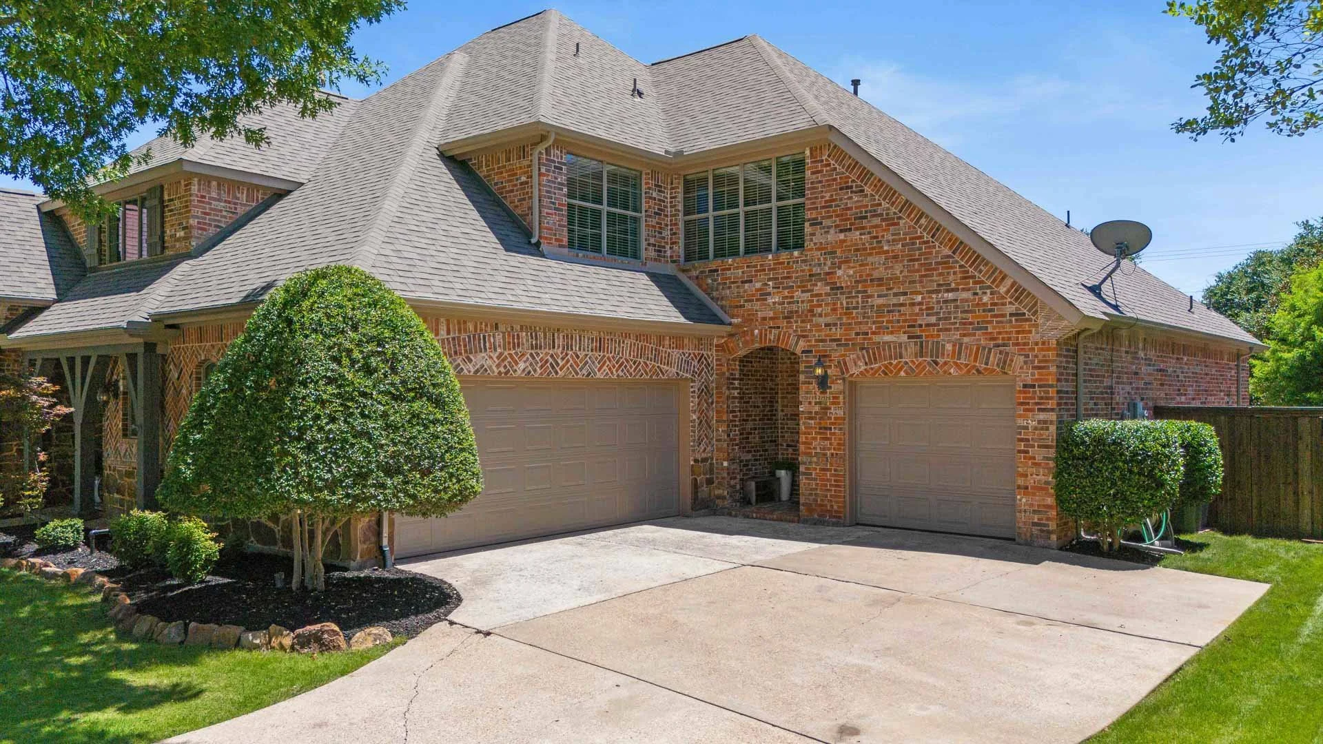 A two-story brick house with a gray shingled roof and a three-car garage. There are two trimmed bushes in front of the house, and a small garden bed with dark mulch and rocks around the bushes. The house has multiple windows with white frames, and a 