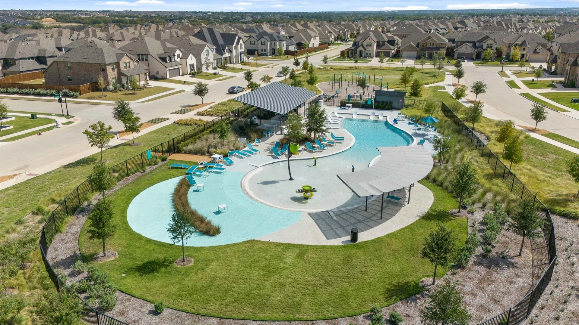 A community swimming pool area surrounded by a black fence, with numerous lounge chairs and umbrellas, in a suburban neighborhood with rows of houses and green lawns.