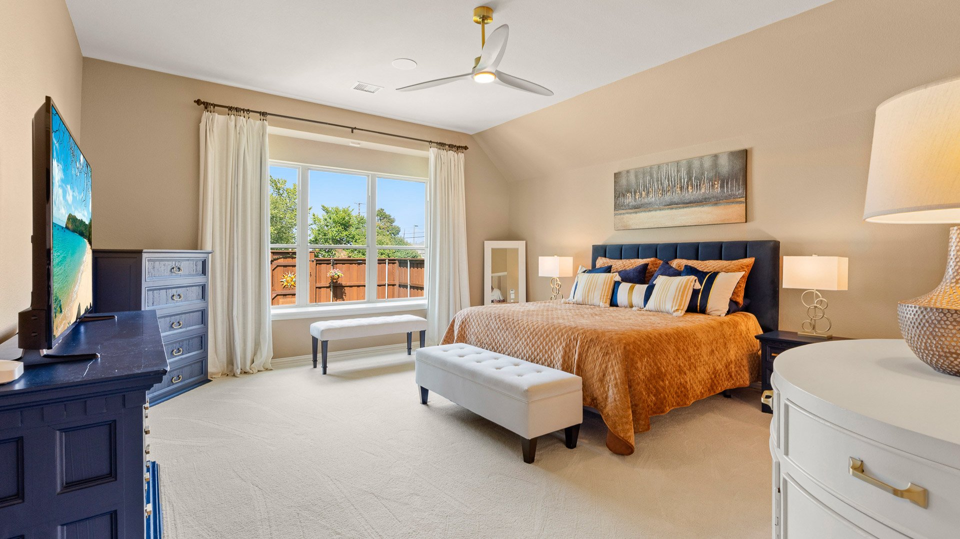 A bedroom with a large bed featuring an orange bedspread and multiple pillows, next to white nightstands with lamps, a white bench at foot of the bed, a sitting window with cream curtains, a dresser with a TV on it, and a ceiling fan, with neutral-co