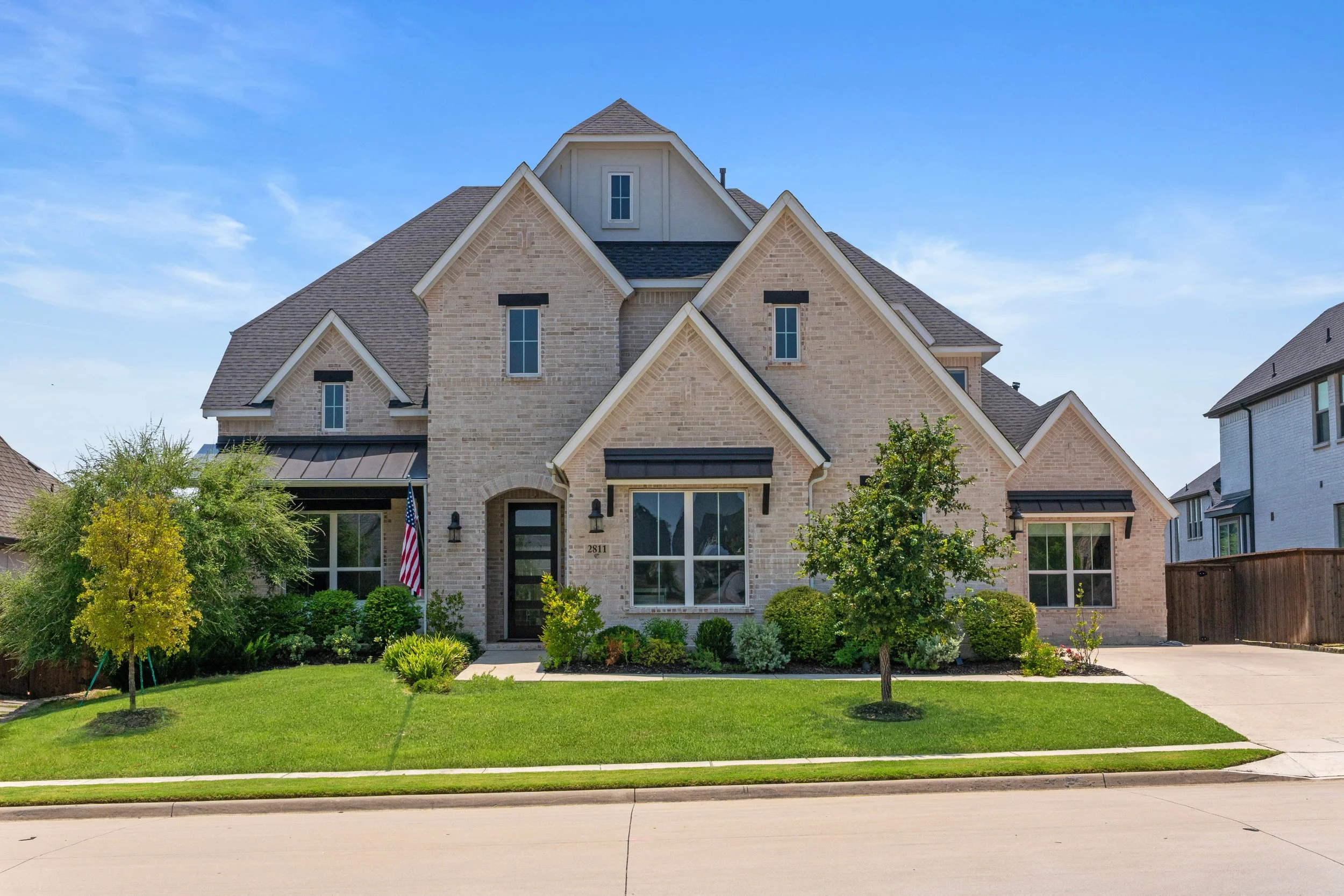A modern, multi-story suburban house with a brick exterior, front lawn, and driveway on a sunny day.