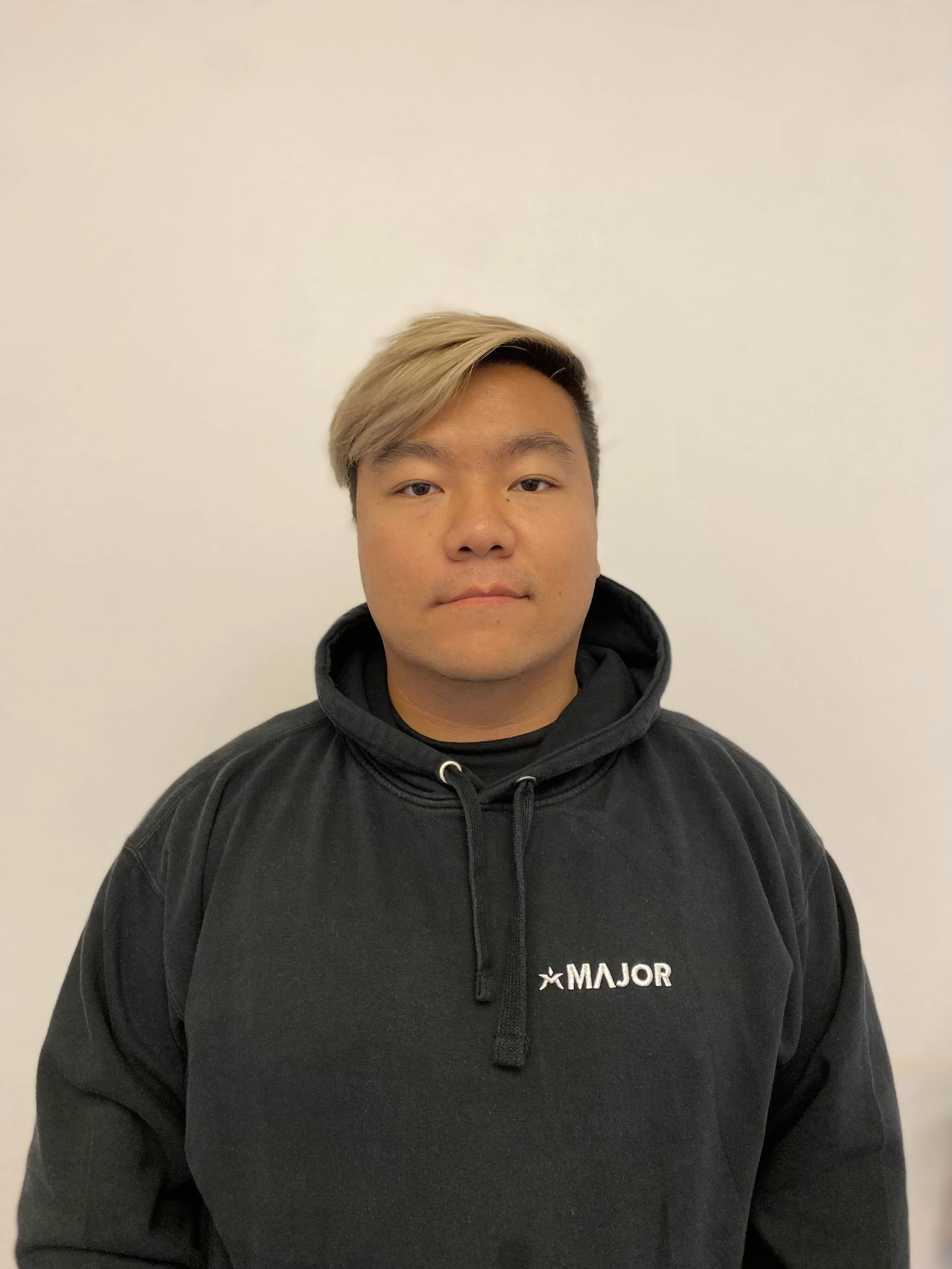 Ung mann /  Datamannen / ITmann / IT-mann / Data mann med blond hår og sort hettegenser står foran en hvit vegg.