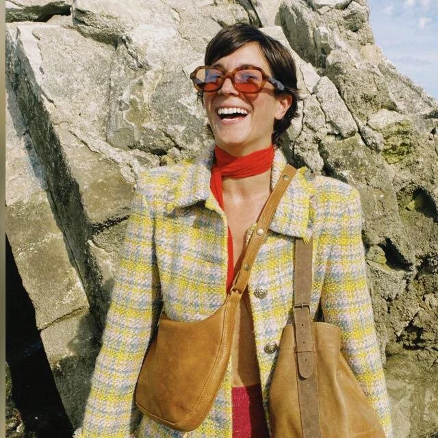 Une femme souriante prend un selfie à l'intérieur. En arrière-plan, une grande fenêtre montre un paysage avec des arbres, une vieille voiture bleue stationnée dans l'herbe, et d'autres véhicules garés dehors.
