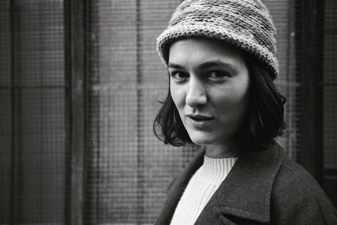 Portrait d'une femme avec un bonnet en tricot, portant un pull blanc et un manteau sombre, devant une grille ou un grillage en arrière-plan.