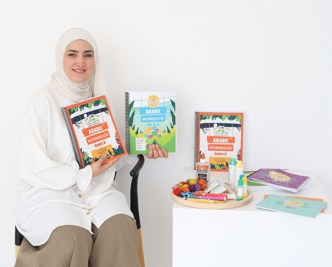 Iman Hasan dressed in modest clothing, sitting on a chair and smiling, holding a spiral-bound Arabic workbook. On a nearby table, there are several other workbooks, craft materials, glue, and colorful pens, all related to Arabic language learning.