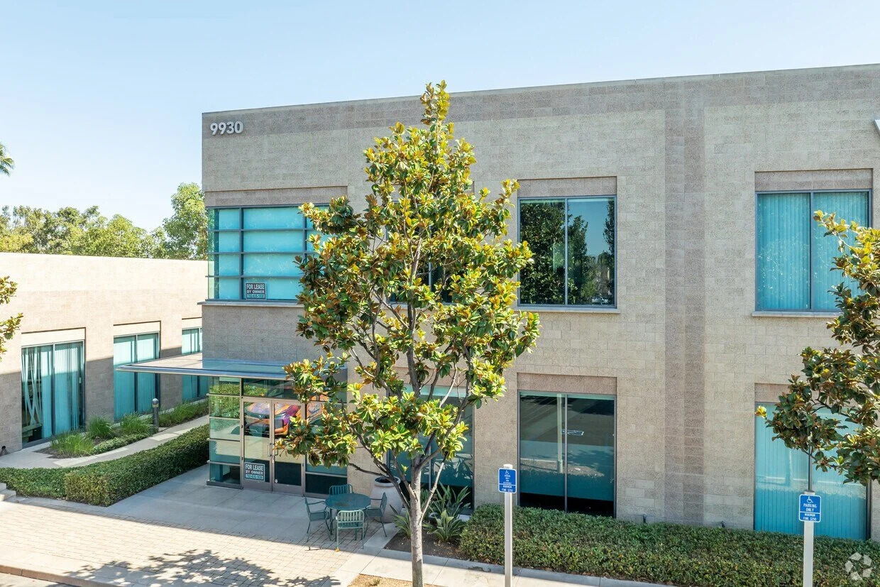 A two-story office building with large windows, trees, and a small parking lot with handicap designated spots.