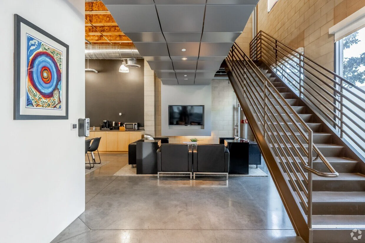 Modern office lounge area with a staircase, black leather chairs, a wall-mounted TV, and a kitchenette in the background.