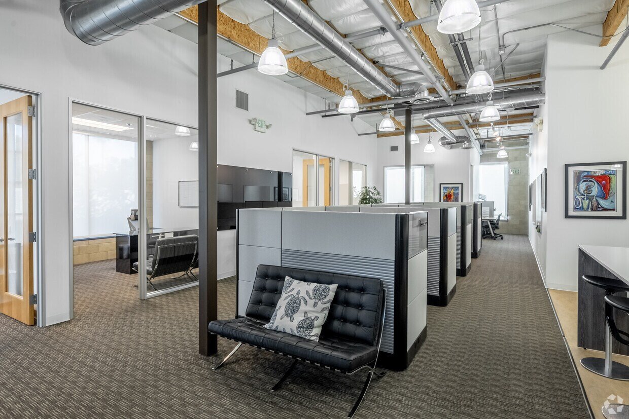 Modern office space with workstations, cubicles, and lounge area with a black sofa and decorative pillow, featuring exposed ceiling pipes and large windows.