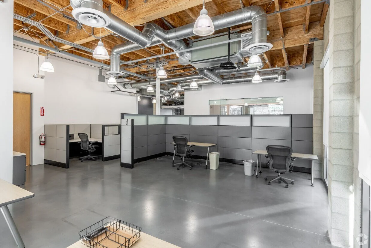 Empty modern office workspace with cubicles, desks, office chairs, and large window, exposed wooden ceiling beams, and visible ductwork.