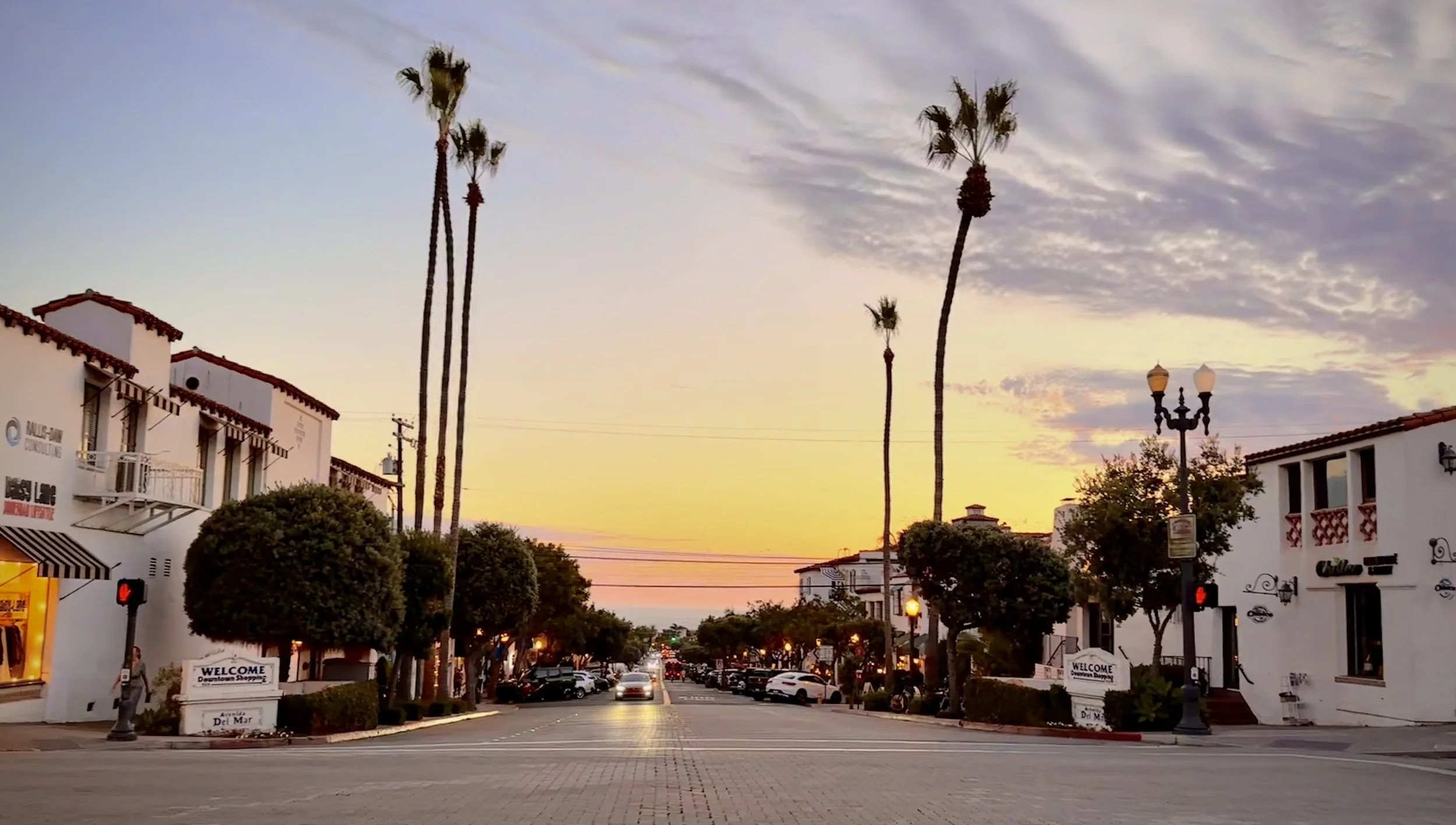 A lively street scene at sunset with palm trees and white buildings, parked cars, streetlights, and a sign that says 'Welcome Downtown Shopping'.