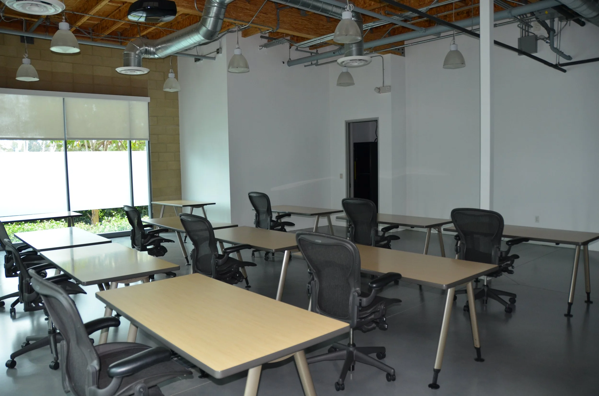 Empty modern conference room with rows of desks and chairs, large window with blinds, exposed ductwork, and white walls.