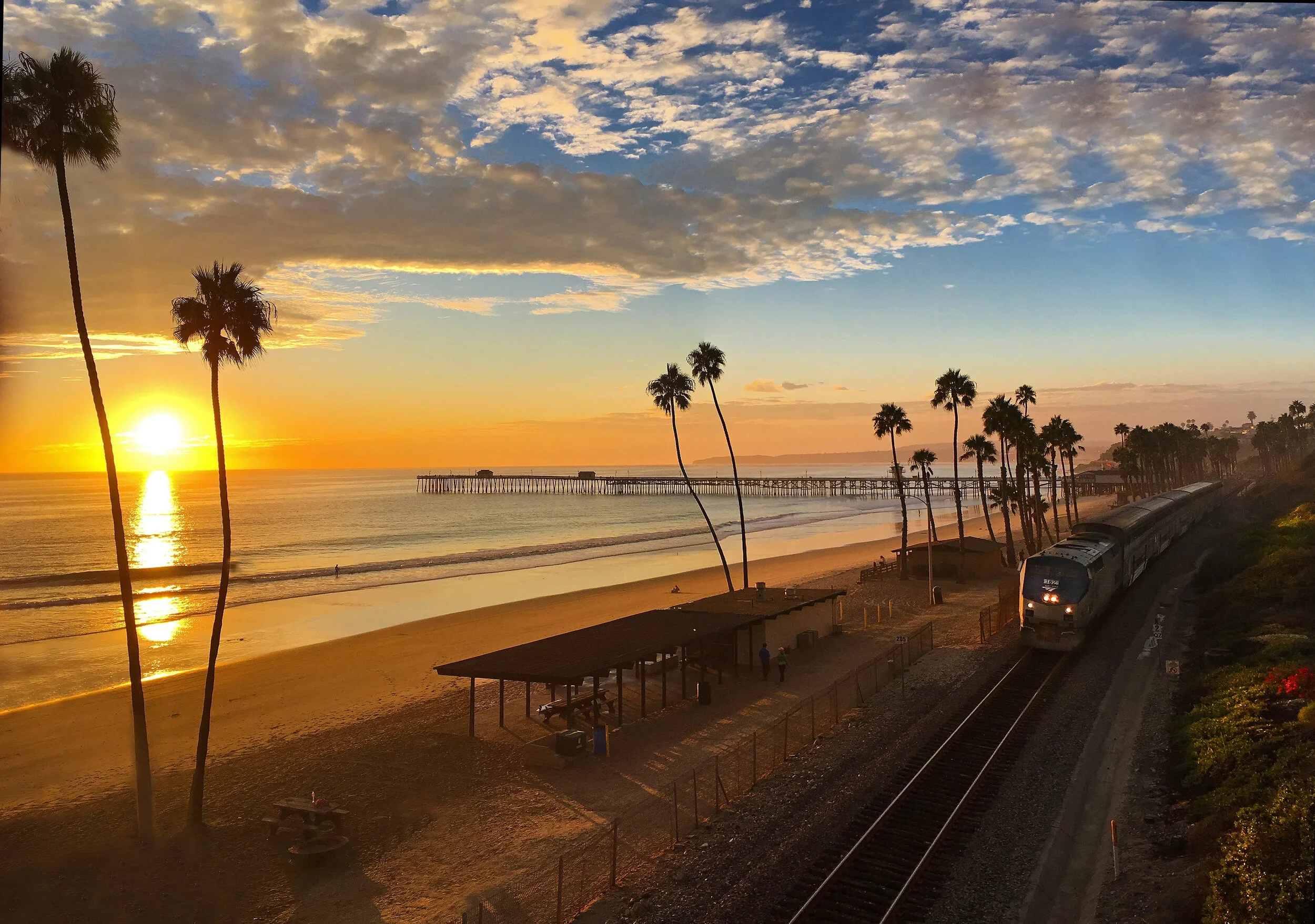 Sunset over the ocean with a pier in the distance, palm trees along the beach, and a train running on tracks near the shoreline.