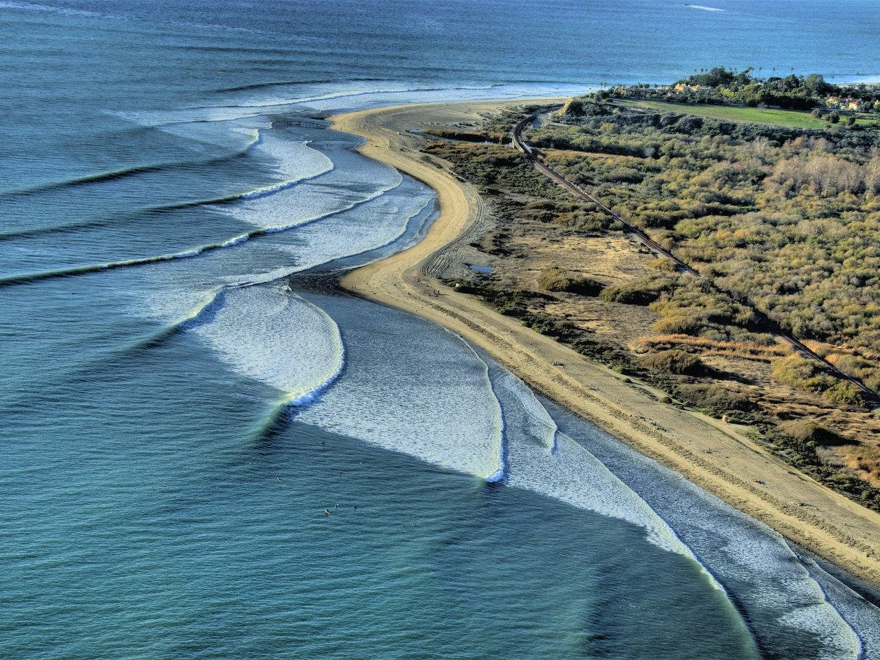 Aerial view of a sandy beach with ocean waves, a shoreline with patches of grass and shrubs, and a railway track running parallel to the coast.