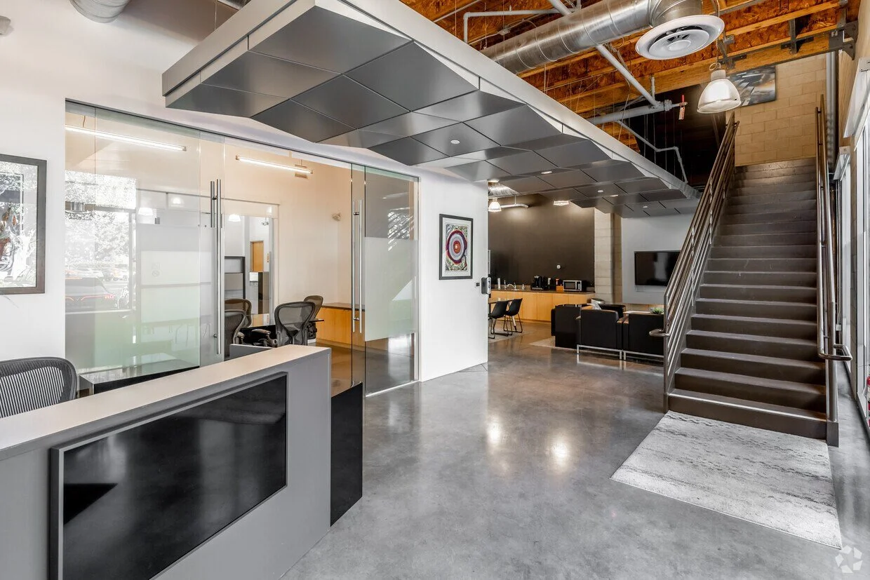 Modern office lobby with a reception desk, glass-walled meeting rooms, a staircase, and a lounge area with chairs and a wall-mounted television.
