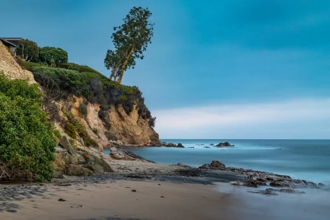A beach with a rocky shoreline and a sandy slope leading up a cliff with trees and vegetation. The ocean extends to the horizon under a cloudy sky.