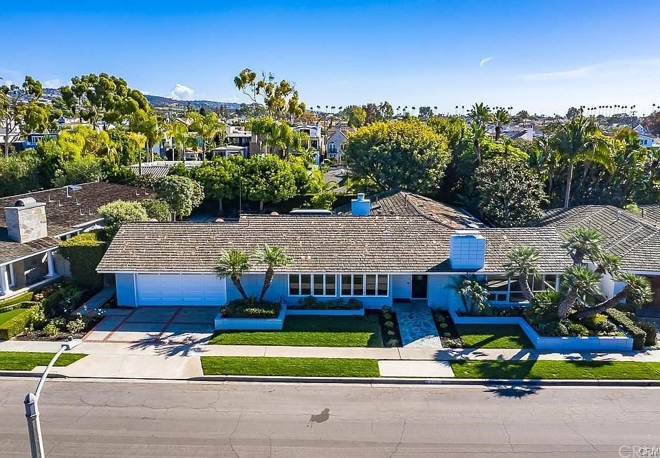 Aerial view of a single-story house with a gray shingle roof, white walls, and a front yard with green grass, palm trees, and flower bushes, situated in a suburban neighborhood with other houses and trees nearby.