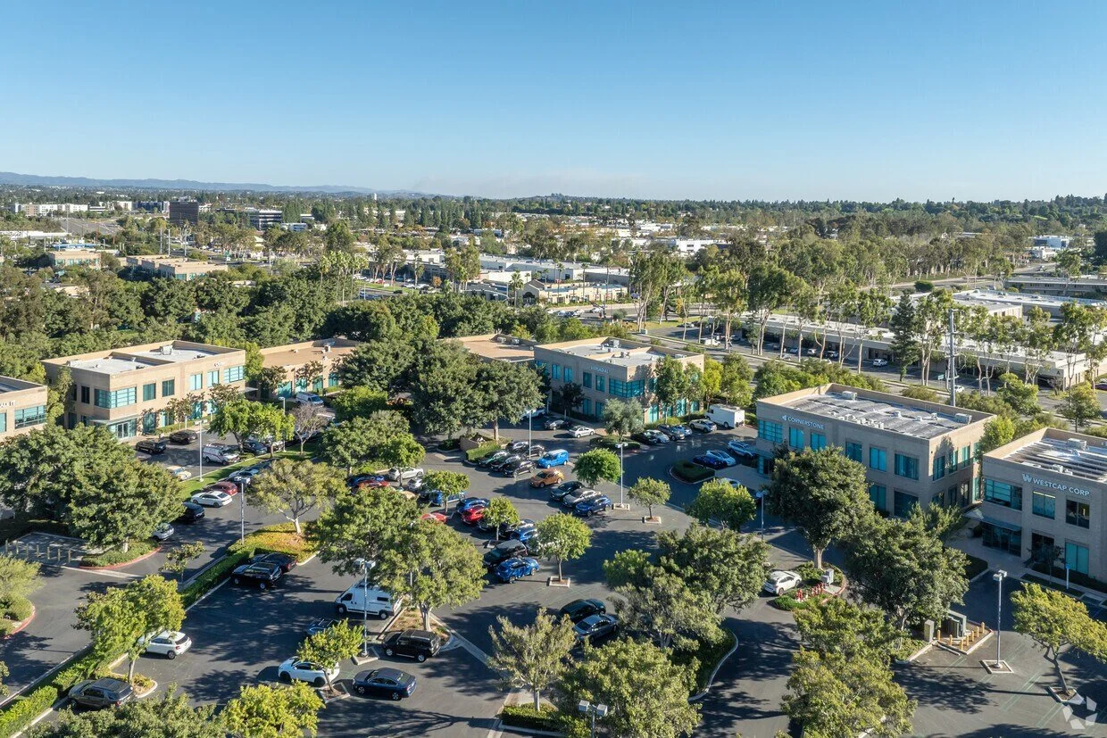 Aerial view of a commercial area with parking lots, office buildings, trees, and roads under a clear blue sky.
