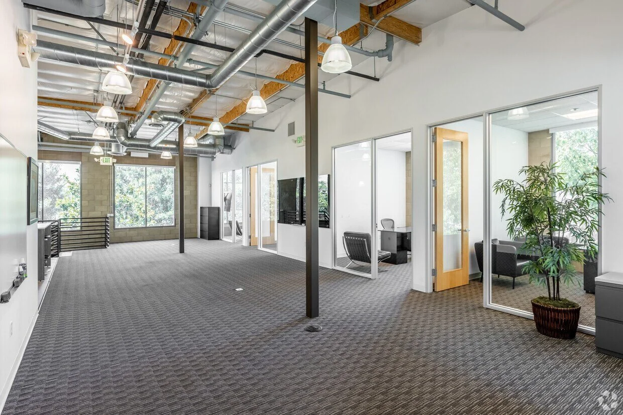 Empty modern office space with large windows, glass-walled meeting rooms, and exposed ceiling pipes, illuminated by hanging lights.