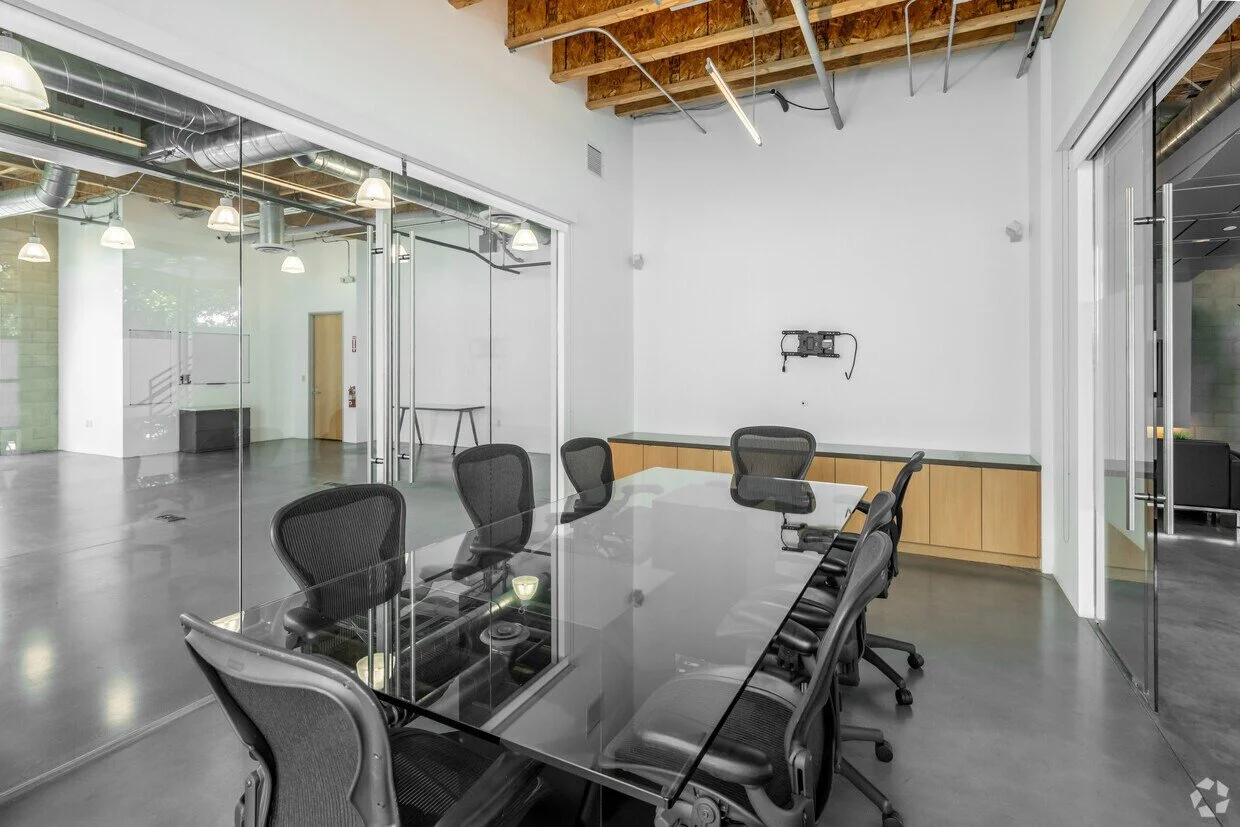 Modern conference room with a glass-top table, black office chairs, and a built-in cabinet along the wall. The room has white walls, a high ceiling with exposed wooden beams, and a glass window wall that reveals an adjacent space with a polished concrete floor and hanging lights.