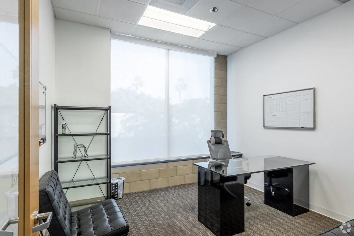 Empty office with a glass table, gray office chair, black sofa, metal shelving unit, whiteboard on the wall, large window with blinds, beige carpet, and ceiling lights.