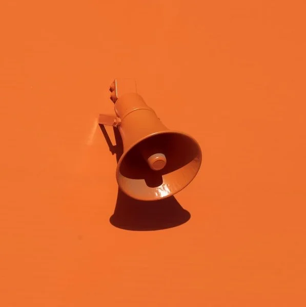 A small gray loudspeaker mounted on an orange wall, casting a shadow.