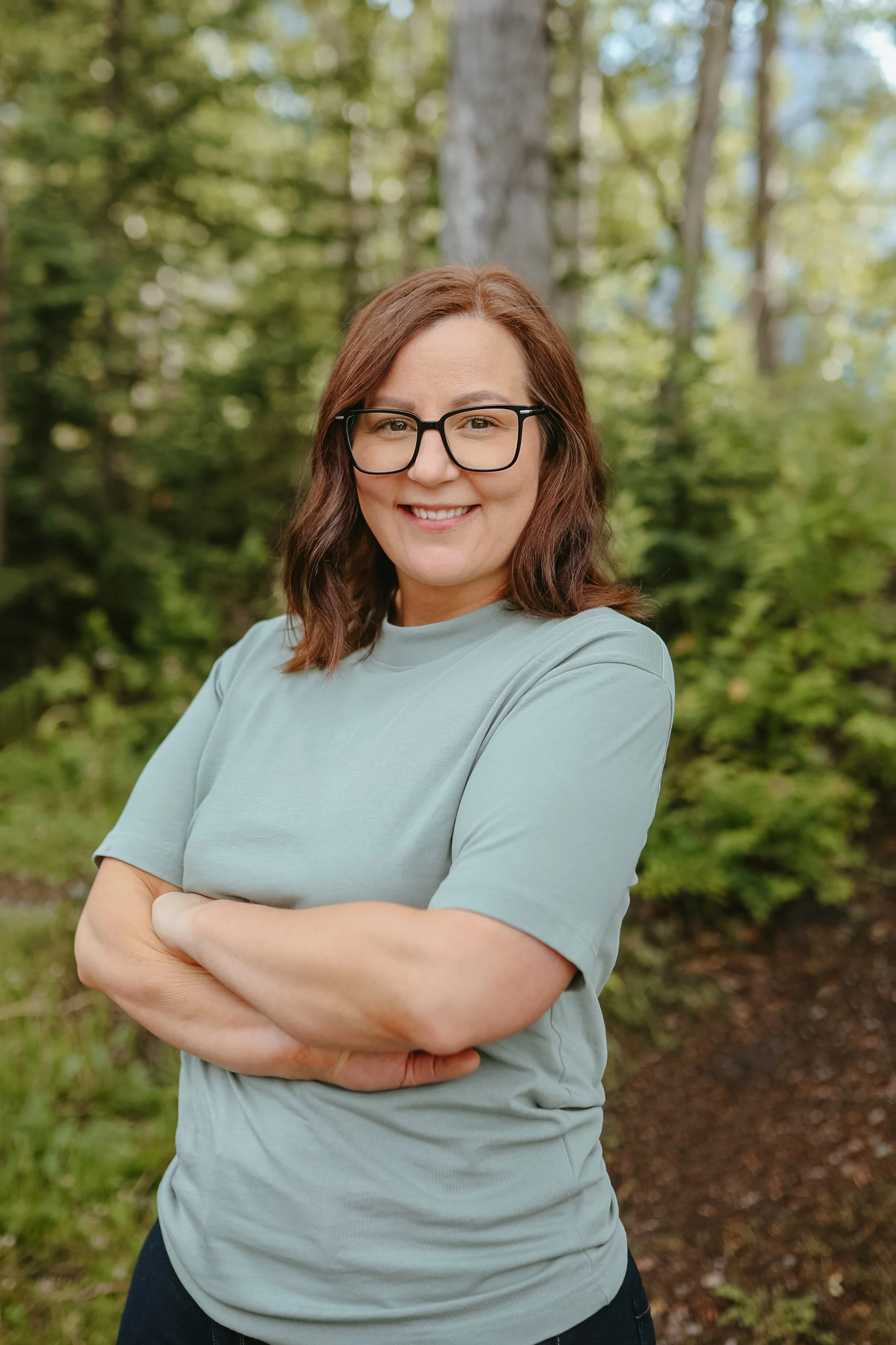 Owner of Sol Havn with shoulder-length brown hair, wearing glasses and a light blue t-shirt, standing outdoors in a wooded area, smiling with arms crossed.
