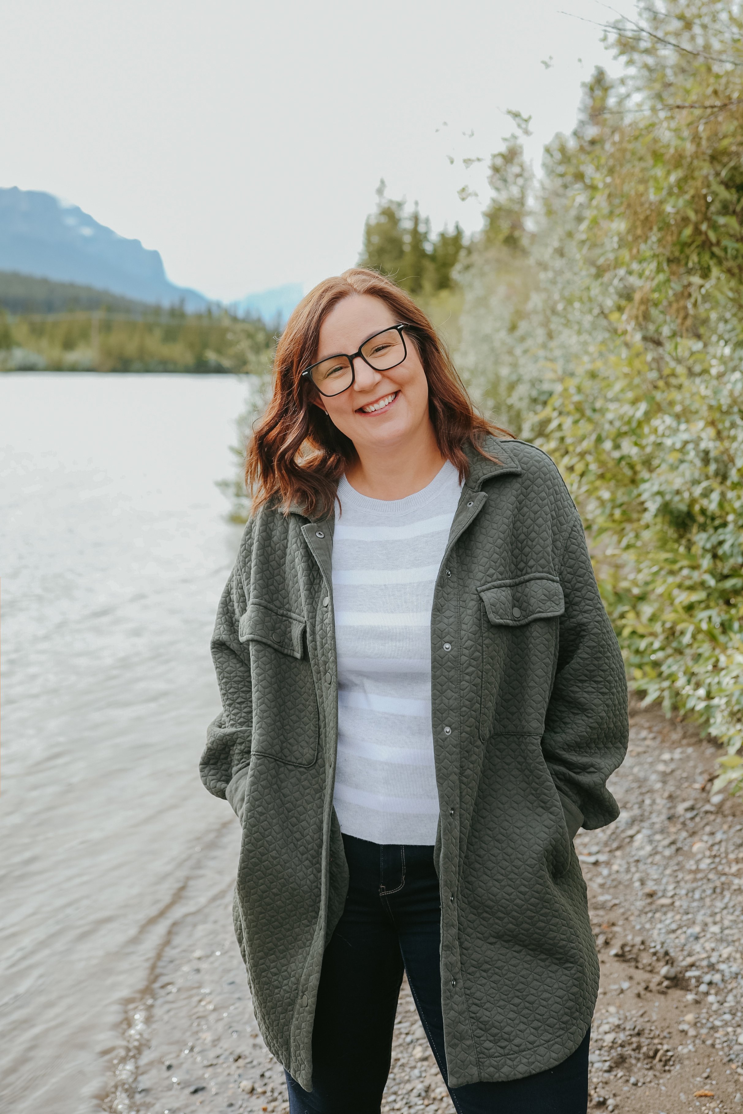 Owner of Sol Havn with glasses smiling standing outdoors near a lake with mountains and trees in the background.