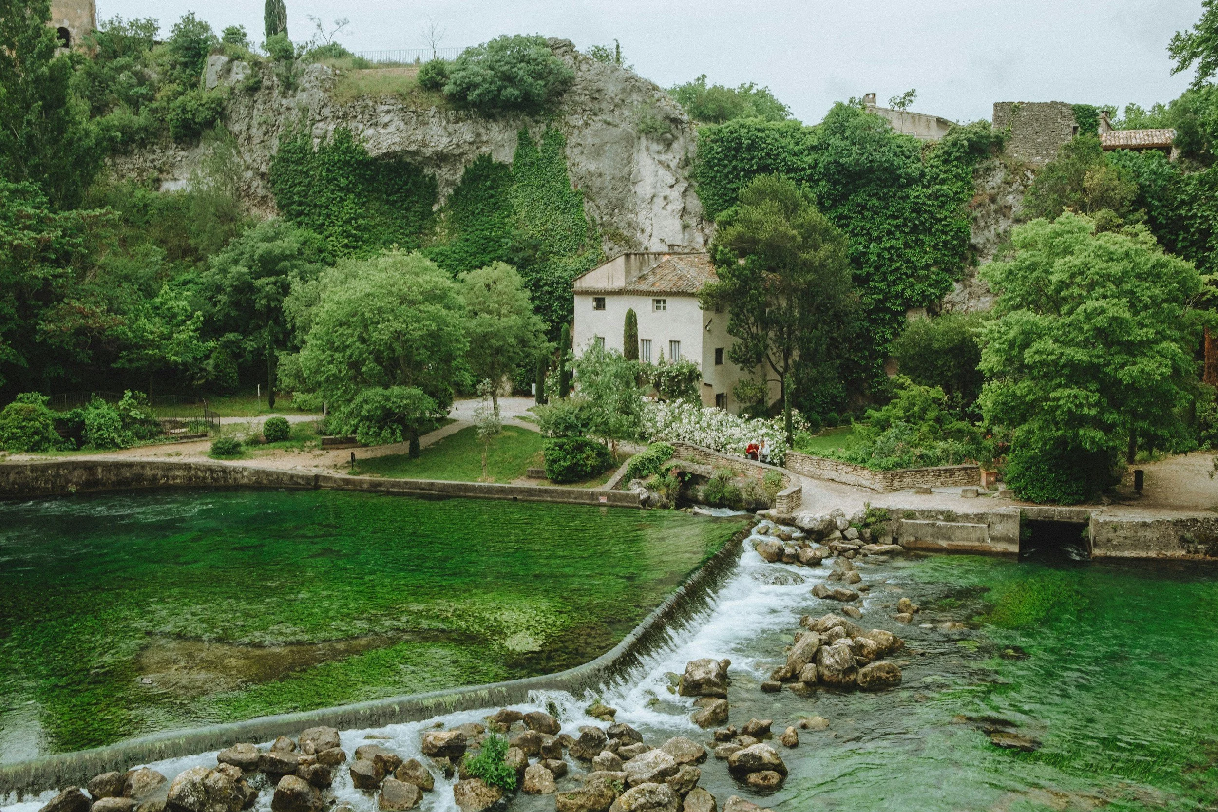 A serene scene of a small house by a river, with lush green trees and bushes surrounding it, and rocky hills in the background.