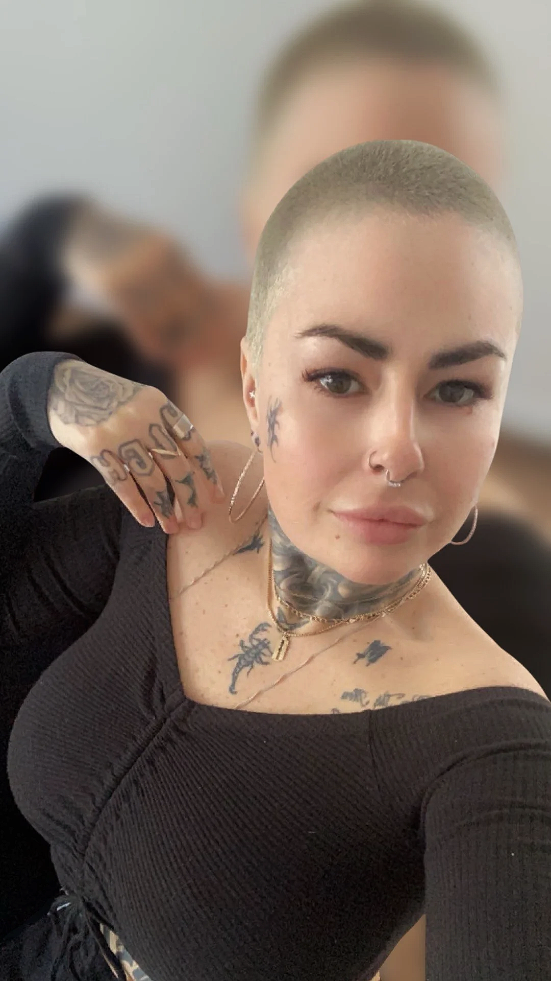 A woman with a shaved head, tattoos, and piercings taking a selfie. She is wearing a black top and jewelry, and has an intense gaze.