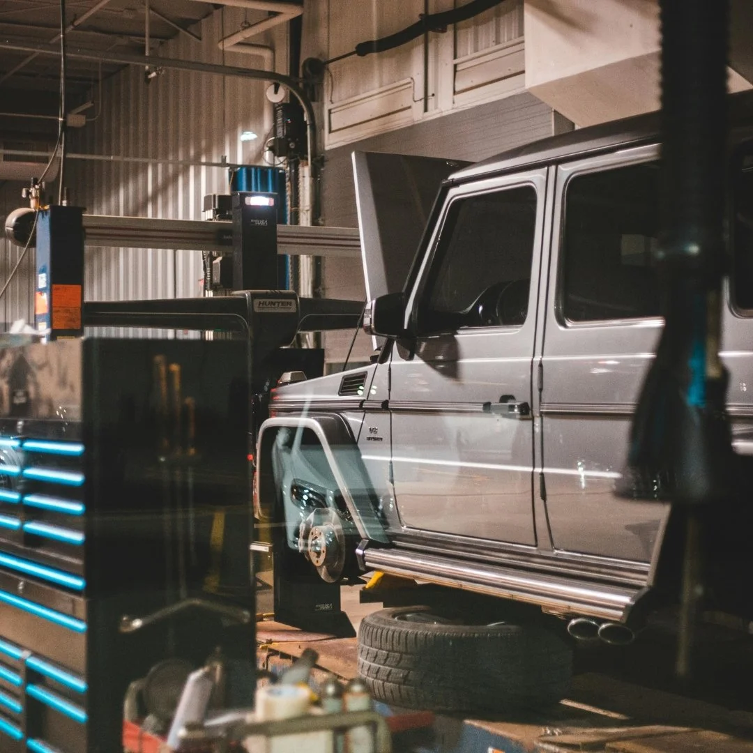 A silver SUV on a lift in a garage or repair shop, with one wheel removed and a tire on the ground below. The garage has tools and equipment around, including a blue tool chest in the foreground.
