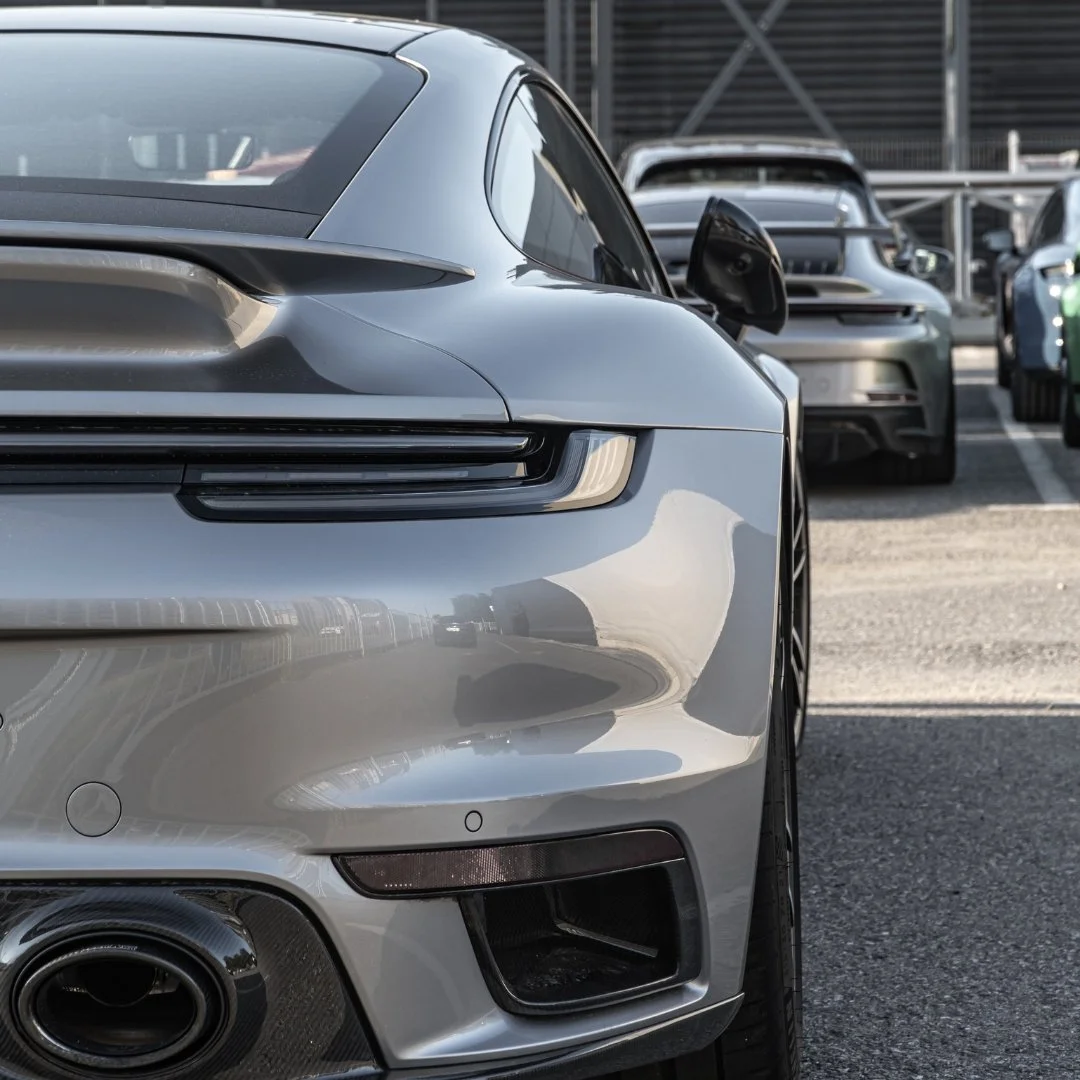 Close-up of the front side of a silver sports car, with a focus on the headlight, side mirror, and wheel, parked in a lot with other cars.