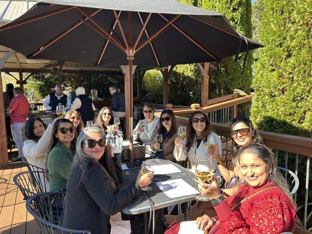 A group of women sitting around a table outdoors, holding wine glasses and smiling, with a patio umbrella overhead. In the background, a bar area and greenery are visible.