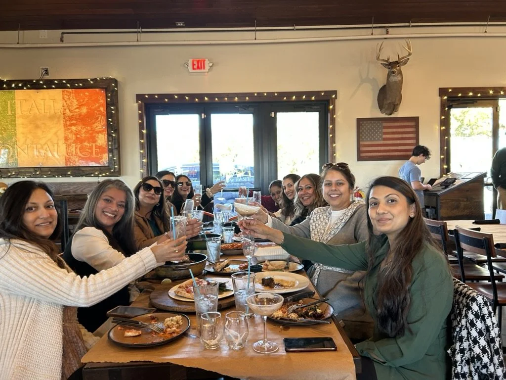 A group of women sitting around a restaurant table, smiling and raising glasses for a toast, with food and drinks in front of them.