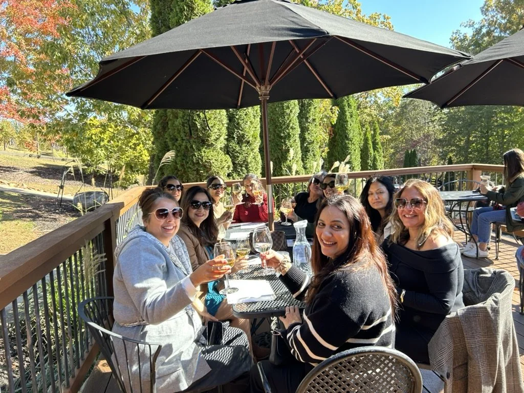 A group of women dining at an outdoor restaurant on a sunny day, sitting under large umbrellas with trees and greenery in the background.