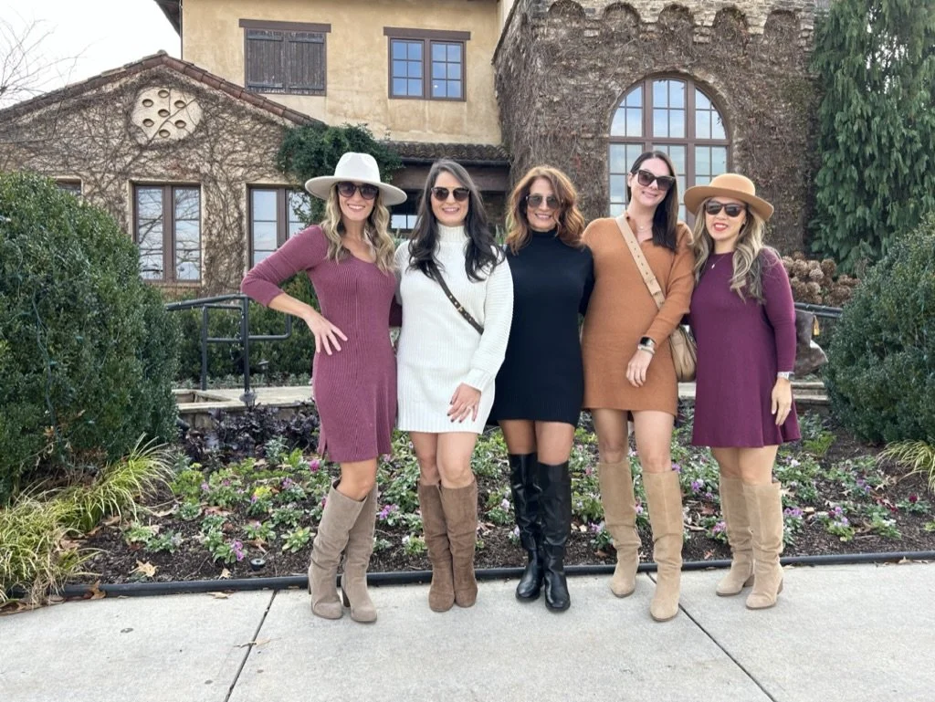 Five women posing outdoors in front of a rustic-style building with greenery and flowerbeds, all wearing stylish fall outfits with boots and sunglasses.