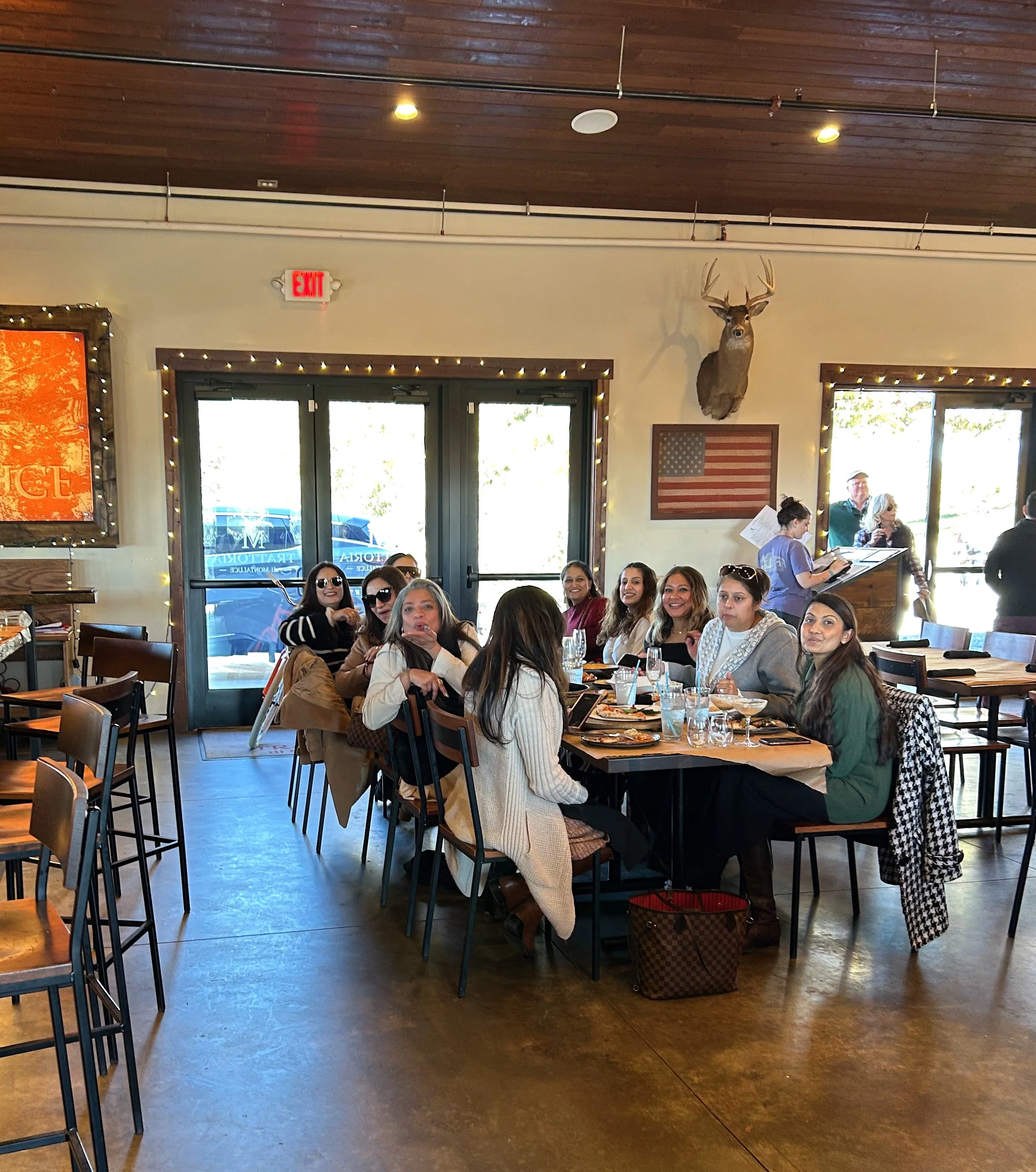 A group of women sitting around a wooden table inside a restaurant, smiling and enjoying food and drinks; a deer head and American flag decor on the wall.