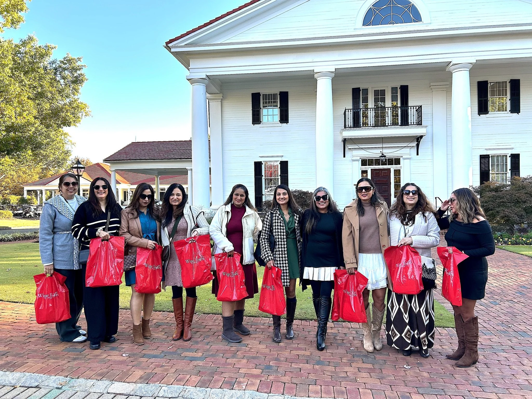 Group of ten women standing on a brick pathway in front of a large white house with Greek columns, holding red shopping bags, all wearing sunglasses.