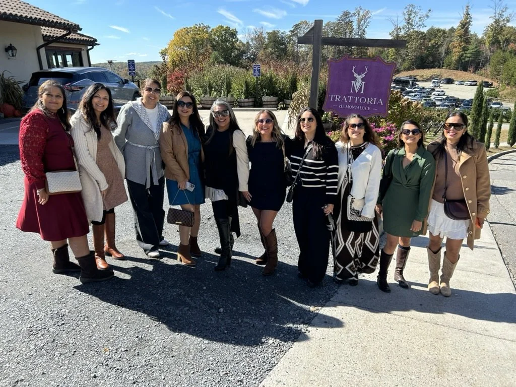 Group of ten women standing outside in front of a sign that reads 'TRATTORIA di MONTALUCE'. They are dressed in fall clothing, smiling, and posing for the photo on a sunny day.