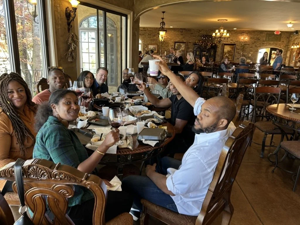 A group of people enjoying a meal at a restaurant, sitting around a large table with food and drinks, some raising glasses for a toast, in a warmly lit dining area with large windows and brick walls.