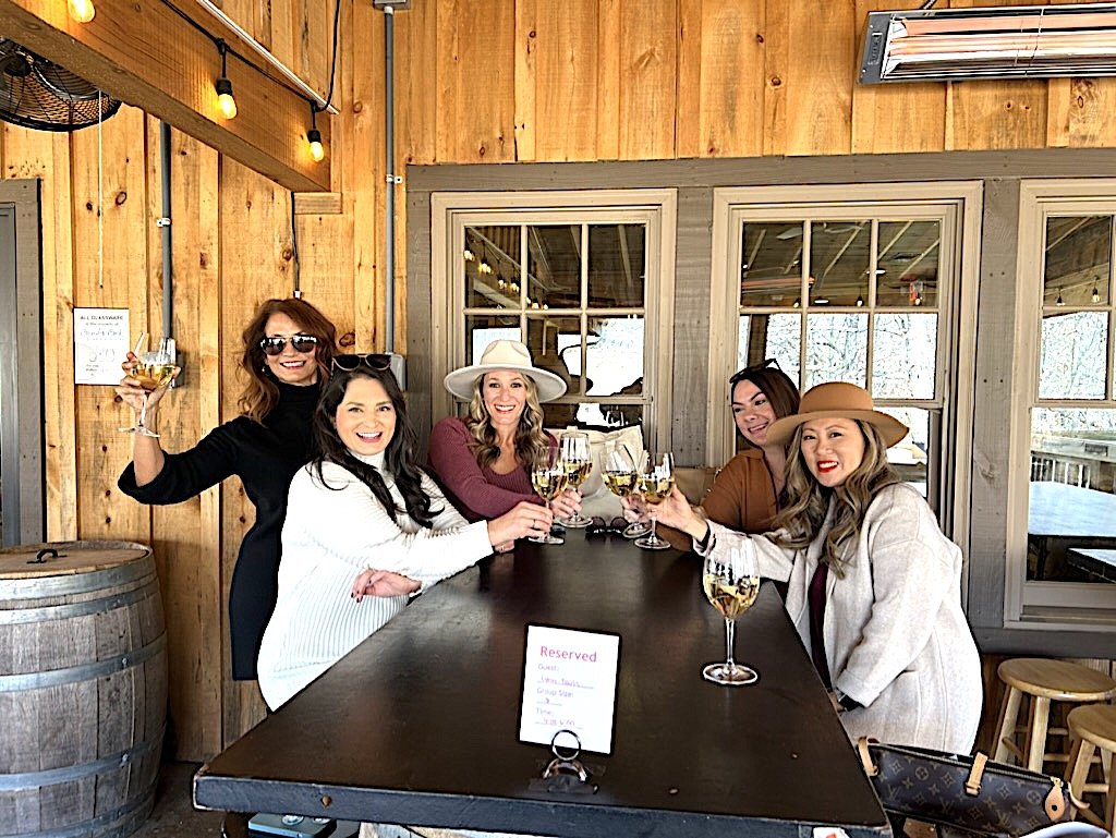 Five women enjoying glasses of white wine at a rustic restaurant. They are smiling and raising their glasses in a toast, sitting around a dark wooden table with a reserved sign. The background features wood-paneled walls, windows, and warm lighting.