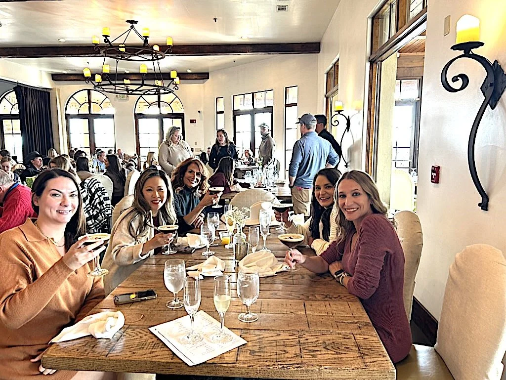 Five women smiling and having drinks at a long dining table in a restaurant, with more people in the background.