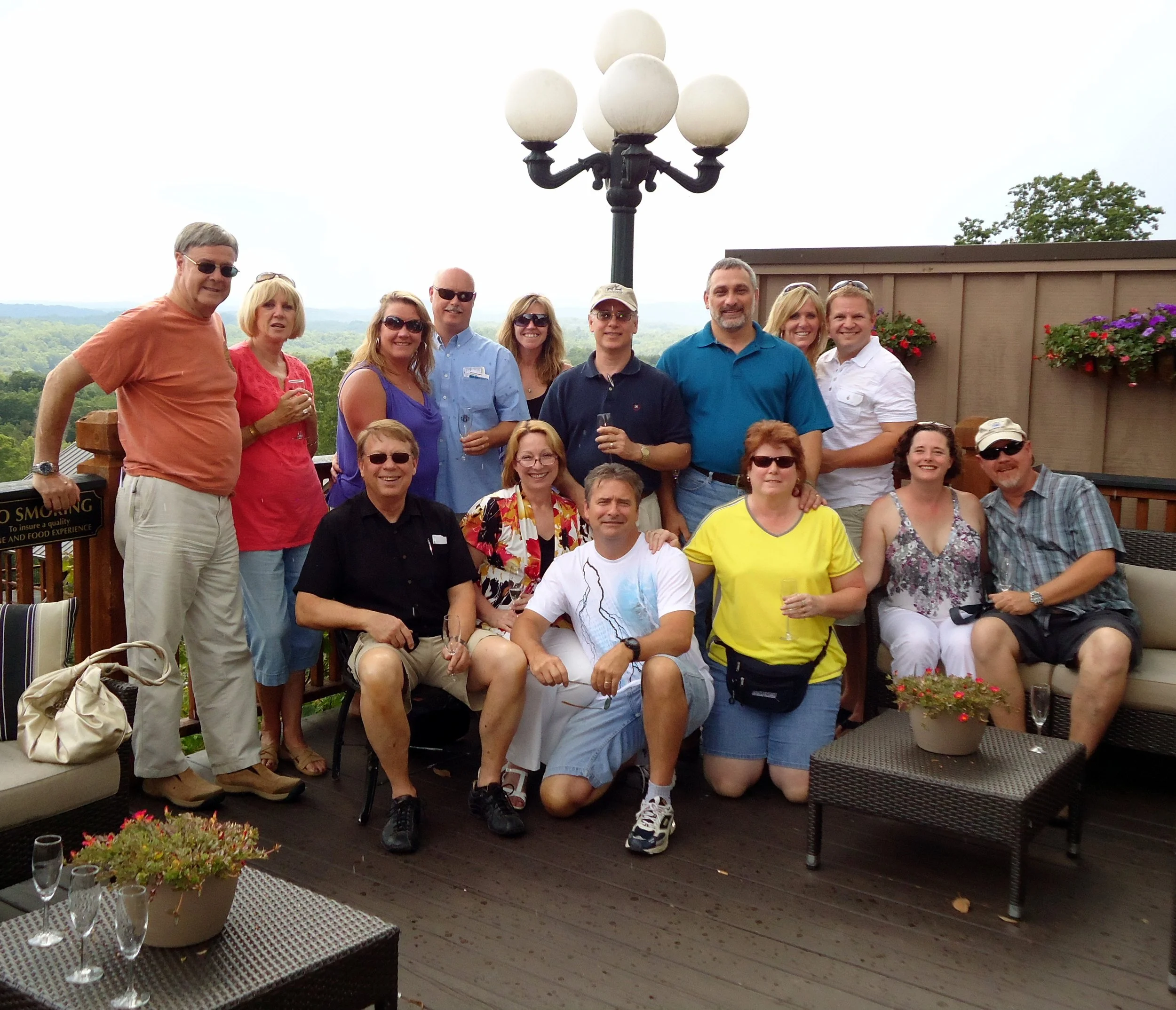 Group of people posing for a photo on a terrace with a scenic view, some holding drinks, with potted plants and outdoor furniture.