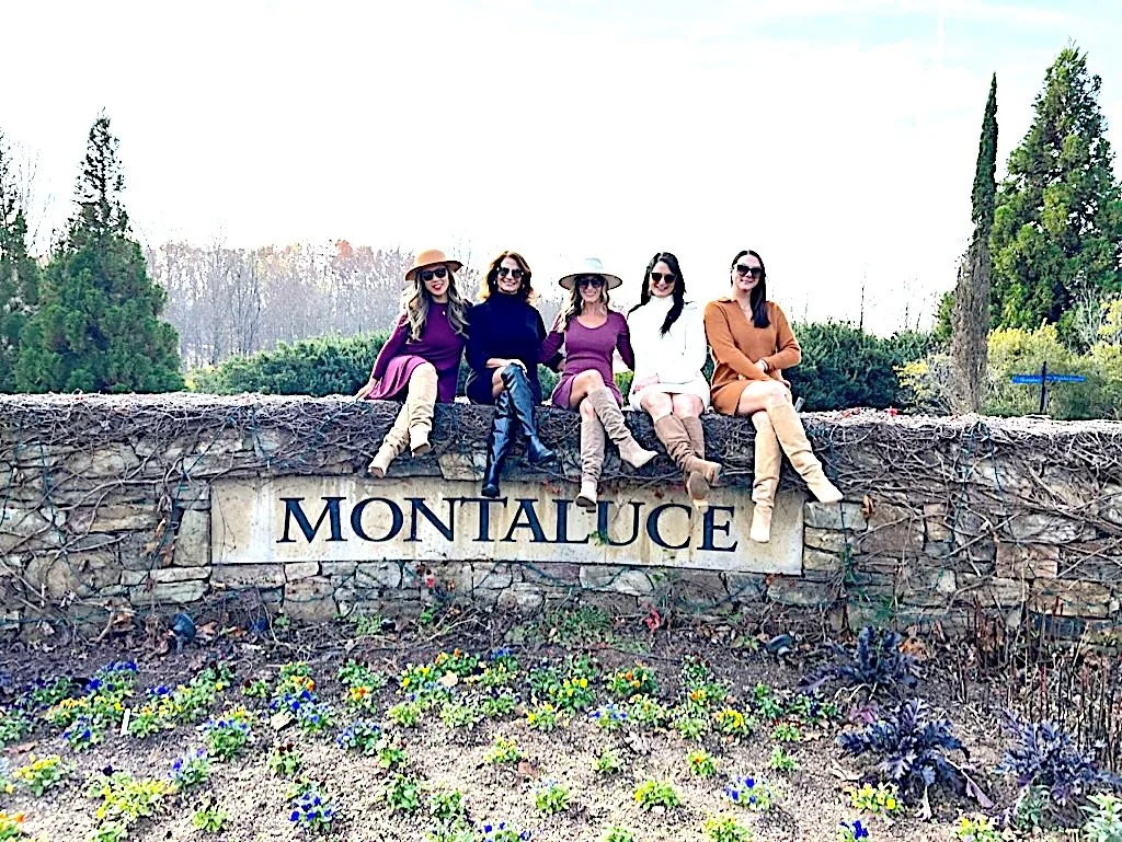 Five women sitting on a stone wall with a sign that reads 'MONTALUCE,' surrounded by plants and trees.