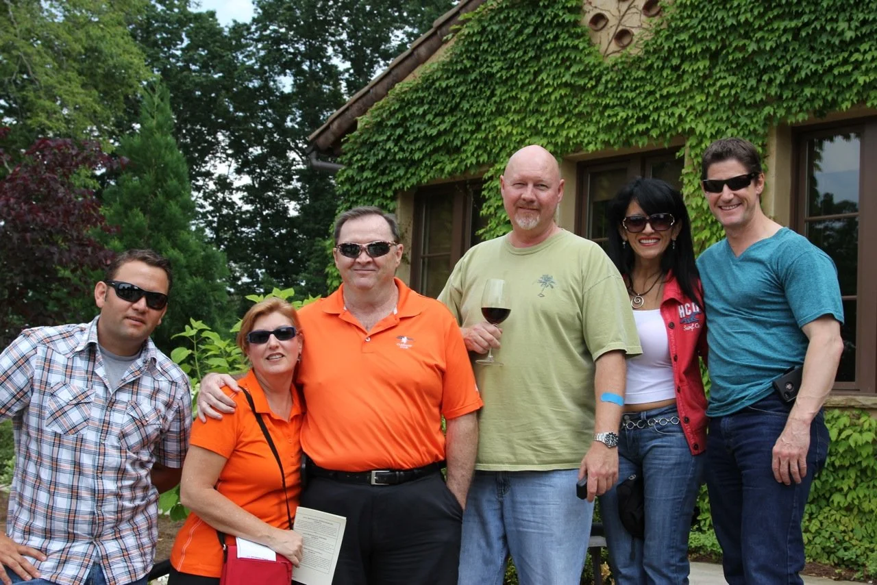 Group of six adults standing outdoors in front of house covered in green ivy, posing for photo, some wearing sunglasses, one holding a glass of red wine.
