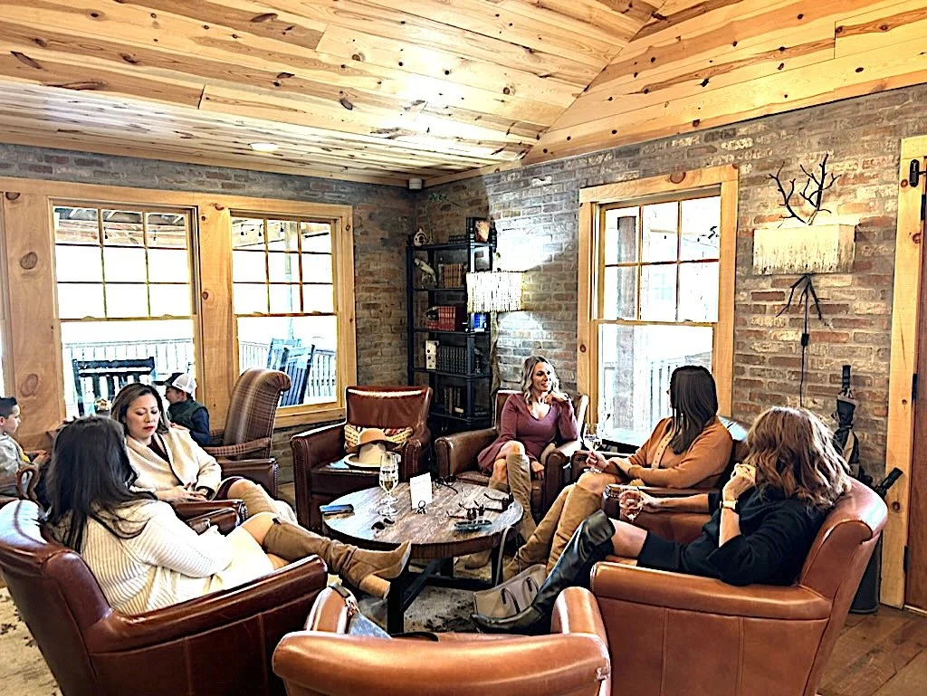 Group of six women sitting and chatting in a cozy rustic living room with large windows, brick walls, leather armchairs, and wooden ceiling.